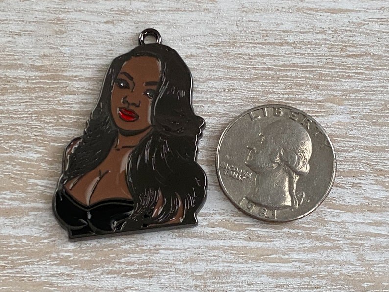 May include: A black metal charm featuring a portrait of a woman with long black hair, wearing a black dress and red lipstick.