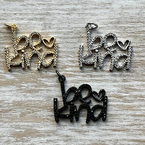 May include: Three charm pendants with the words "be kind" in a cursive font with a small heart. The charms are available in gold, silver, and black.