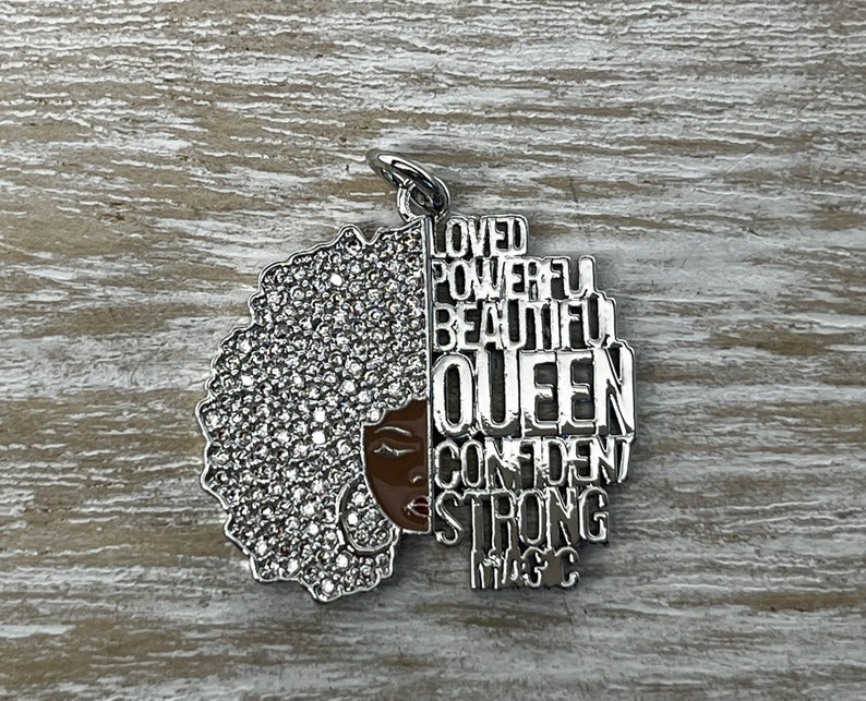 May include: Silver charm with a black woman's face and afro hair with rhinestones. The charm has the words "Loved, Powerful, Beautiful, Queen, Confident, Strong, Magic" around the face.