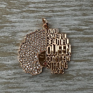 May include: Gold-tone pendant featuring a stylized silhouette of a woman with an afro, adorned with clear rhinestones. The silhouette is outlined with the words "Loved", "Powerful", "Beautiful", "Queen", "Confident", "Strong", and "Magic".