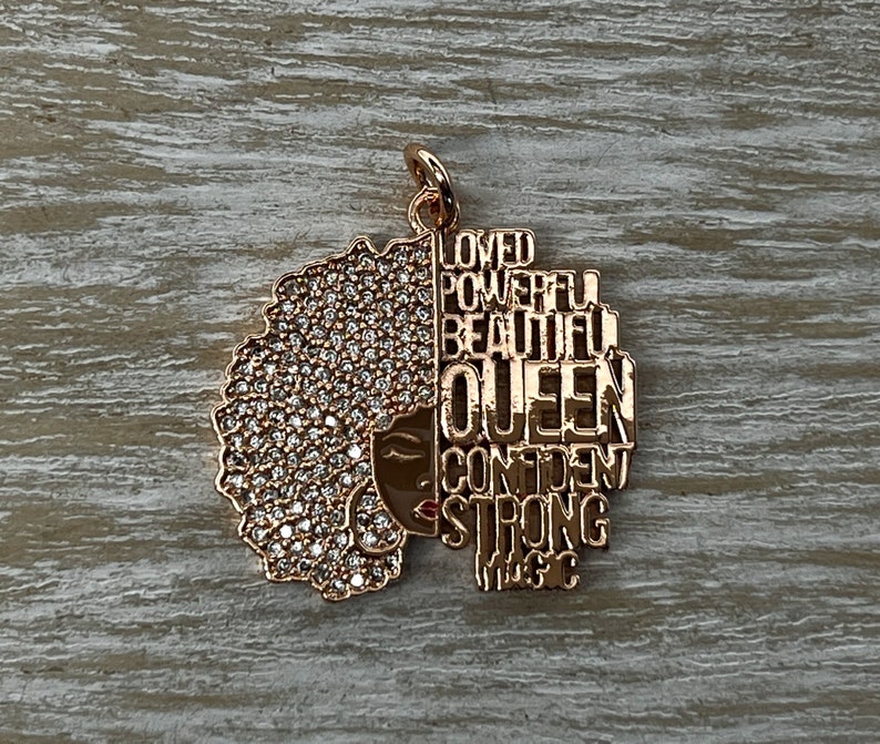 May include: Gold-tone pendant featuring a stylized silhouette of a woman with an afro, adorned with clear rhinestones. The silhouette is outlined with the words "Loved", "Powerful", "Beautiful", "Queen", "Confident", "Strong", and "Magic".