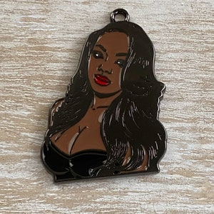 May include: Black metal charm featuring a portrait of a woman with long black hair and red lips. The charm has a shiny finish.