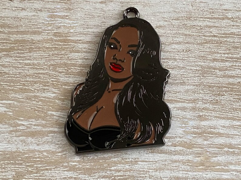 May include: Black metal charm featuring a portrait of a woman with long black hair and red lips. The charm has a shiny finish.