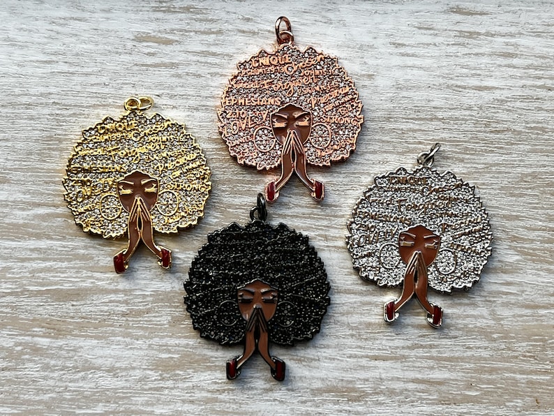 May include: Four metal charms featuring a woman with her hands clasped in prayer. The charms are in gold, silver, black, and rose gold. Each charm has a different design of words around the woman's head.