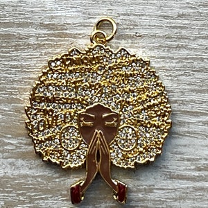 May include: Gold pendant featuring a stylized image of a woman with a large afro hairstyle covered in rhinestones. The woman is depicted with her hands clasped in prayer.