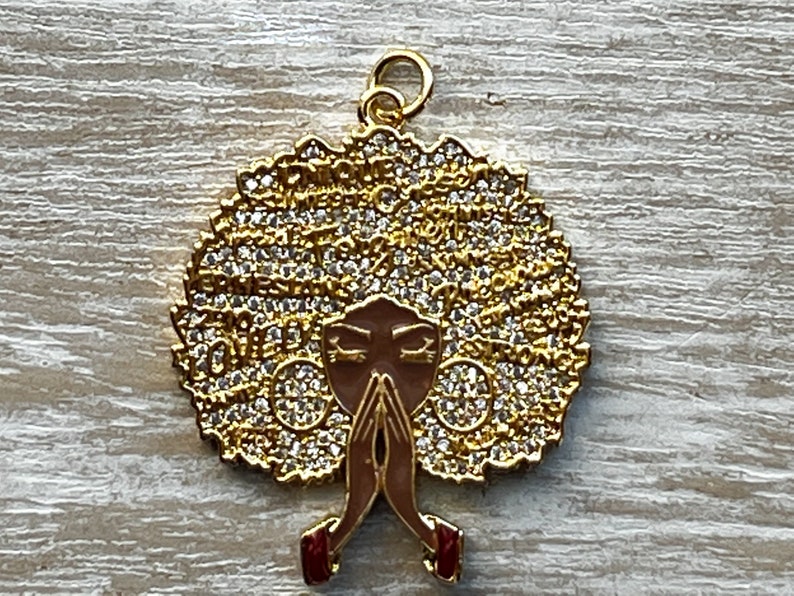 May include: Gold pendant featuring a stylized image of a woman with a large afro hairstyle covered in rhinestones. The woman is depicted with her hands clasped in prayer.