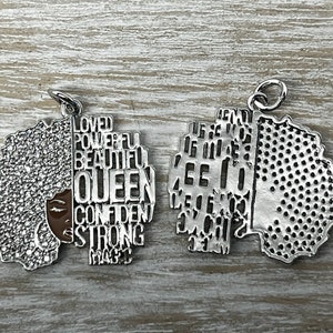 May include: Silver charm with a detailed outline of a woman's face with an afro hairstyle. The charm is divided in half, with one side featuring the words "Loved, Powerful, Beautiful, Queen, Confident, Strong, Magic" and the other side featuring a textured pattern.