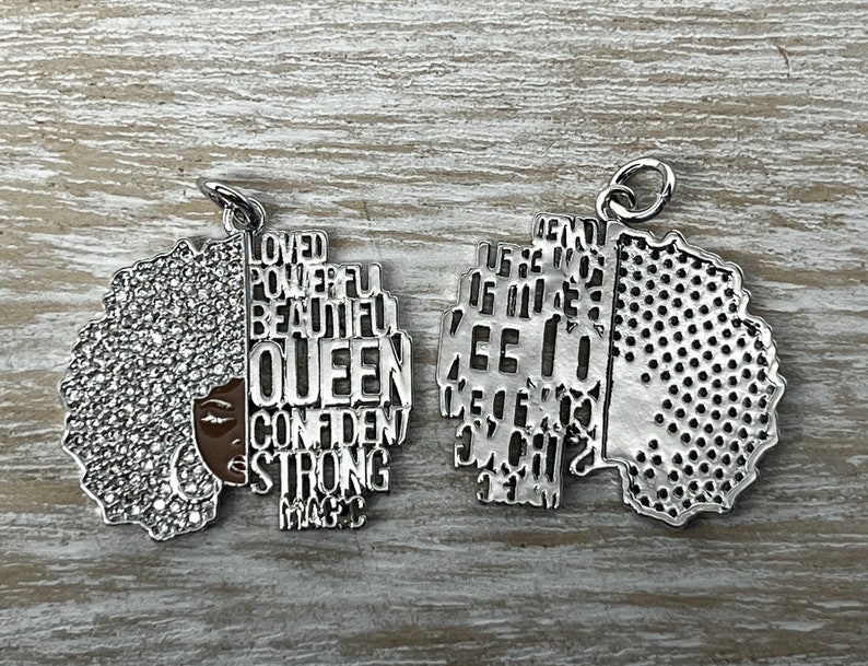 May include: Silver charm with a detailed outline of a woman's face with an afro hairstyle. The charm is divided in half, with one side featuring the words "Loved, Powerful, Beautiful, Queen, Confident, Strong, Magic" and the other side featuring a textured pattern.