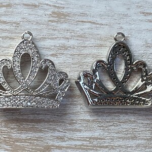 May include: Two silver crown charms with clear rhinestone accents. The crowns are detailed with intricate designs and are perfect for adding a touch of royalty to jewelry projects.