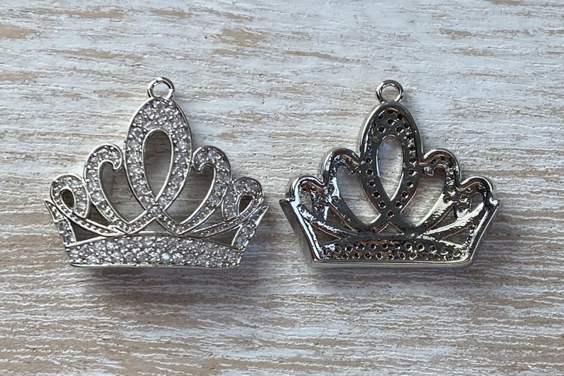 May include: Two silver crown charms with clear rhinestone accents. The crowns are detailed with intricate designs and are perfect for adding a touch of royalty to jewelry projects.