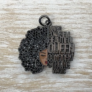 May include: Black metal charm with a silhouette of a woman with an afro hairstyle. The afro is made of black rhinestones. The words "Loved, Powerful, Beautiful, Queen, Confident, Strong, Magic" are written around the silhouette.
