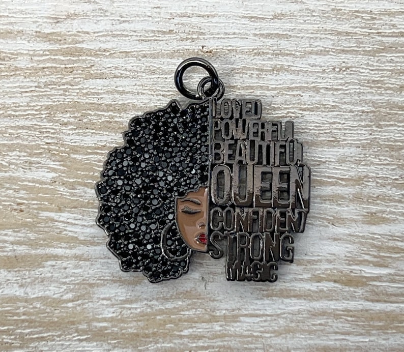 May include: Black metal charm with a silhouette of a woman with an afro hairstyle. The afro is made of black rhinestones. The words "Loved, Powerful, Beautiful, Queen, Confident, Strong, Magic" are written around the silhouette.