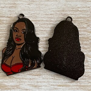 May include: A metal charm featuring a woman with long black hair wearing a red top. The charm is shown with the front and back sides.