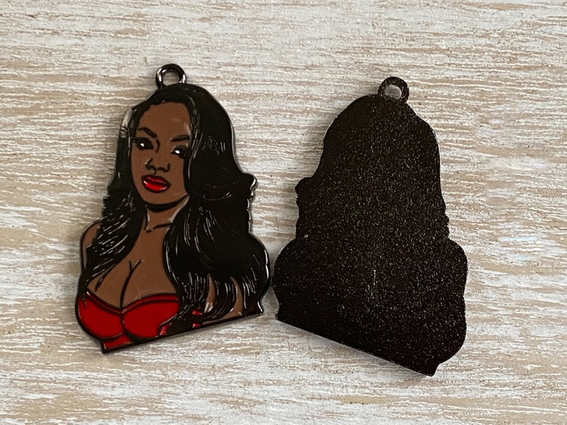 May include: A metal charm featuring a woman with long black hair wearing a red top. The charm is shown with the front and back sides.