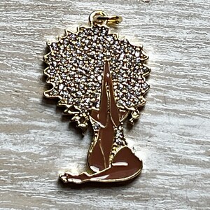 Pode incluir: Encanto de ouro com uma mulher com um grande afro coberto de strass, sentada com as m&atilde;os juntas em ora&ccedil;&atilde;o. A mulher &eacute; morena e o afro &eacute; dourado.