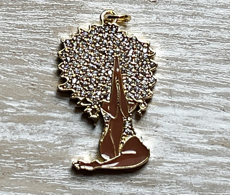 Pode incluir: Encanto de ouro com uma mulher com um grande afro coberto de strass, sentada com as m&atilde;os juntas em ora&ccedil;&atilde;o. A mulher &eacute; morena e o afro &eacute; dourado.