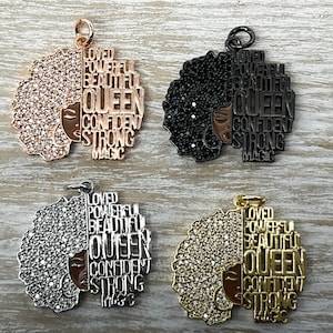 May include: Four gold-tone, silver-tone, and black charms featuring a woman's face with an afro hairstyle. The charms are adorned with rhinestones and the words "Loved", "Powerful", "Beautiful", "Queen", "Confident", "Strong", and "Magic" are inscribed on the charms.