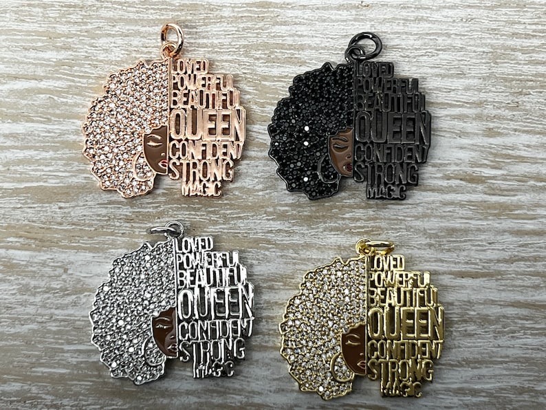 May include: Four gold-tone, silver-tone, and black charms featuring a woman's face with an afro hairstyle. The charms are adorned with rhinestones and the words "Loved", "Powerful", "Beautiful", "Queen", "Confident", "Strong", and "Magic" are inscribed on the charms.