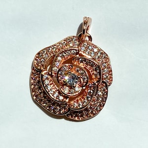 May include: A rose-gold pendant featuring a rose design with clear cubic zirconia stones. The pendant has a loop at the top for attaching to a chain.