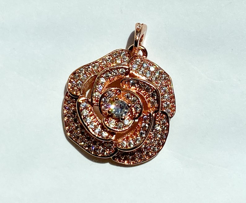 May include: A rose-gold pendant featuring a rose design with clear cubic zirconia stones. The pendant has a loop at the top for attaching to a chain.