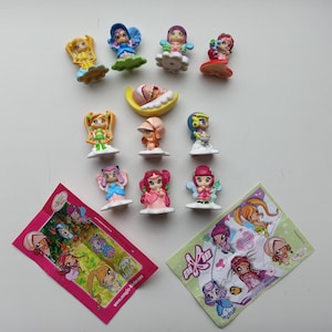 Pode incluir: Uma coleção de dez pequenas figuras de plástico coloridas com vários personagens de olhos grandes e trajes distintos, acompanhadas por dois encartes de papel retangulares com gráficos e o texto Magic Kinder e Pixie.