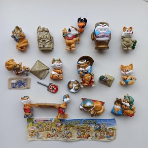 Puede incluir: Una colección de figuras "Miezi Cats" de Kinder Sorpresa. Las figuras son de varios tonos de marrón, naranja y blanco, algunas con sombreros y sosteniendo objetos. Una pancarta en la parte inferior muestra el logotipo de "Miezi Cats" e ilustraciones de personajes.