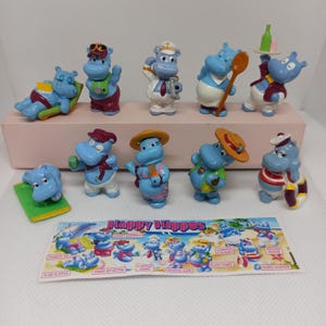 Könnte beinhalten: Eine Sammlung blauer Happy Hippos-Figuren aus Kunststoff in verschiedenen Posen und Outfits. Einige Flusspferde tragen Hüte, Kochjacken und Matrosenuniformen. Ein farbenfroher Happy Hippos-Aufkleber mit Cartoon-Flusspferden ist ebenfalls vorhanden.