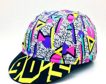 May include: A black and yellow cycling cap with a geometric pattern in pink, purple, blue, and turquoise. The cap has a white background with a black and white geometric pattern.