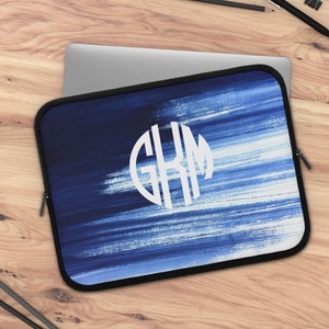 Puede incluir: Una funda para portátil azul y blanca con un monograma blanco "GKM" en un círculo. La funda tiene una cremallera.