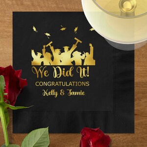 May include: Black graduation party napkin with gold text and graphics. The text reads "We Did It! Congratulations Kelly & Jamie" with silhouettes of graduates. A red rose and a glass of wine are in the image.