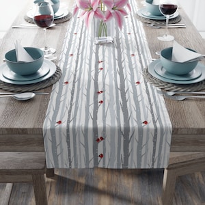 Puede incluir: Un camino de mesa blanco con un diseño de bosque de invierno. El camino de mesa presenta árboles grises con puntos blancos y cardenales rojos. El camino de mesa está sobre una mesa de madera con cubiertos.