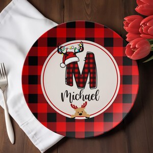 May include: A round Christmas plate with a red and black buffalo plaid pattern. The center features a white circle with a plaid "M" and the name "Michael". A reindeer and Santa hat decorate the plate.