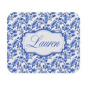 May include: A square coaster with a white background and a blue floral pattern. The center features a white label with the name "Lauren" in blue script, framed by a blue border. The coaster has rounded corners.