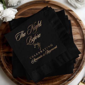 May include: A stack of black cocktail napkins with gold lettering that reads "The Night Before" and "Celebrating Amanda & Raymond". The napkins have a ribbed border and are arranged on a wooden tray.