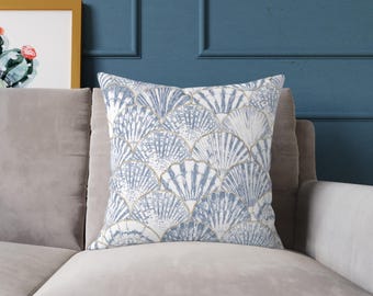 Pode incluir: Almofada quadrada com um padrão de conchas azuis e brancas. A almofada tem uma borda dourada e está colocada em um sofá cinza claro. O fundo é uma parede azul com um quadro emoldurado.