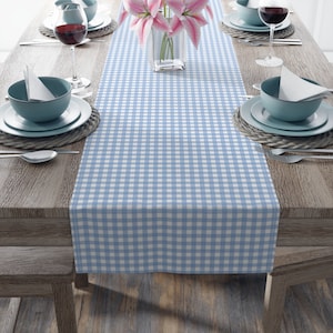 May include: A blue and white gingham table runner with a checkered pattern. The runner is laid out on a wooden table with place settings on either side.