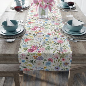 May include: A white table runner with a floral pattern featuring pink, blue, yellow, and green flowers. The runner is on a wooden table with two place settings on each side.