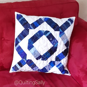 Könnte beinhalten: Ein weißes Kissen mit einem blauen und weißen geometrischen Muster. Das Kissen liegt auf einem roten Sofa. Der Text "@quiltingSally" befindet sich auf dem Sofa.