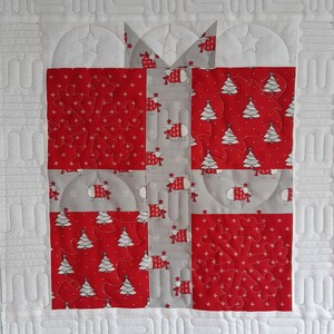 May include: A quilted fabric gift box with a red and white snowflake design, a red and white Christmas tree design, and a gray and white sheep design. The box is surrounded by a white border.