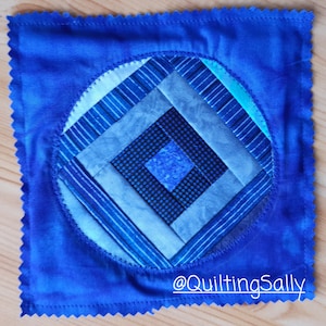 Könnte beinhalten: Ein blauer gesteppter quadratischer Stoff mit einem runden Design, das ein Diamantenmuster in Blautönen zeigt. Die Mitte des Diamanten ist schwarz mit weißen Punkten. Der Text "@quiltingSally" befindet sich in weiß am unteren Rand des Quadrats.