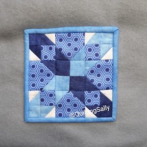 Könnte beinhalten: Ein blauer und weißer Patchwork-Quiltblock mit einem Sternmuster. Der Block ist von einem blauen Rand umgeben. Der Text "@QuiltingSally" befindet sich in der unteren rechten Ecke.