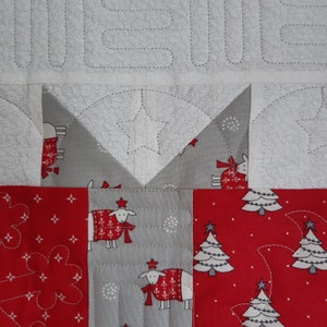 May include: A close-up of a quilt block with a red, white, and gray patchwork design. The block features a star, a Christmas tree, and a sheep with a red scarf.