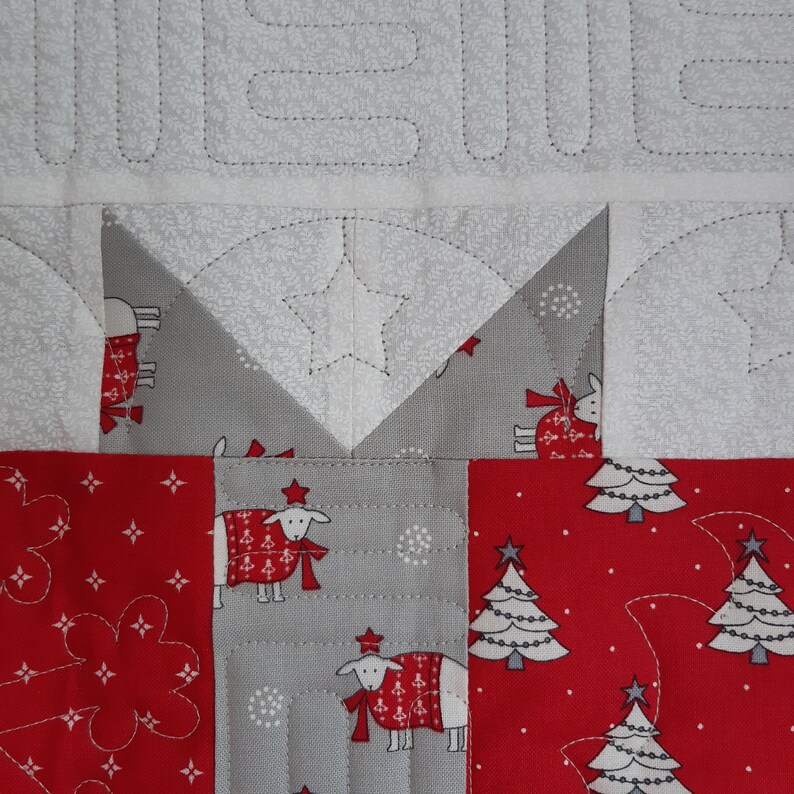 May include: A close-up of a quilt block with a red, white, and gray patchwork design. The block features a star, a Christmas tree, and a sheep with a red scarf.