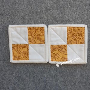 Könnte beinhalten: Zwei weiße und gelbe Stoffuntersetzer mit geometrischem Muster. Die Untersetzer sind gesteppt und haben einen weißen Rand. Der Text "@quiltingSally" ist in der rechten unteren Ecke des Bildes sichtbar.