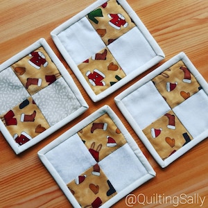Könnte beinhalten: Vier handgefertigte quadratische Untersetzer mit weißem Hintergrund und einem Patchwork-Design aus beigefarbenem Stoff mit rot-weiß-grünen weihnachtlichen Mustern. Die Untersetzer sind mit einem weißen Rand versehen.