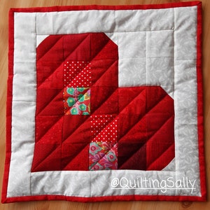 Könnte beinhalten: Eine rote und weiße Patchwork-Decke mit einer Herzform in der Mitte. Das Herz besteht aus verschiedenen roten Stoffen mit weißen Punkten und Blumenmustern. Die Decke ist mit einem roten Rand eingefasst und hat einen weißen Hintergrund mit einem dezenten Muster. Der Text "@QuiltingSally" befindet sich in der rechten unteren Ecke.