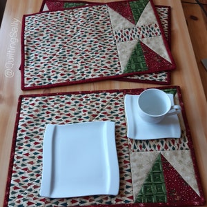 Könnte beinhalten: Zwei gequiltete Tischsets mit einem Weihnachtsbaum-Muster. Die Tischsets sind rot, grün und beige. Ein Tischset hat einen weißen Teller und das andere eine weiße Untertasse und Tasse.