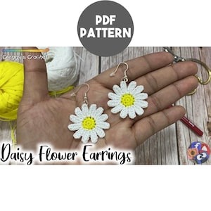 May include: A pair of crochet daisy flower earrings featuring white petals and a bright yellow center, attached to silver-toned metal earring hooks. The image includes the text PDF PATTERN, Creggy's Crochet, and Daisy Flower Earrings.