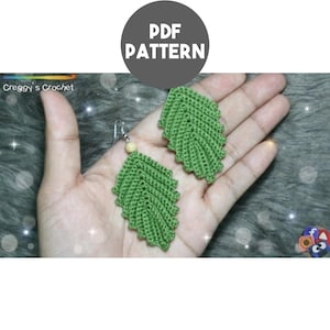 May include: A pair of green crochet leaf earrings featuring a textured stitch pattern and a small light-colored bead at the top of one earring. The image includes the text PDF PATTERN and the logo for Creggy's Crochet in the upper section.