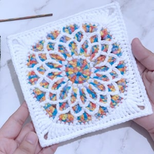 K&ouml;nnte beinhalten: Ein wei&szlig;es H&auml;kelquadrat mit einem bunten Mandala-Design in der Mitte. Das Mandala ist aus verschiedenen Blaut&ouml;nen, Rosa, Gelb und Wei&szlig; gefertigt.